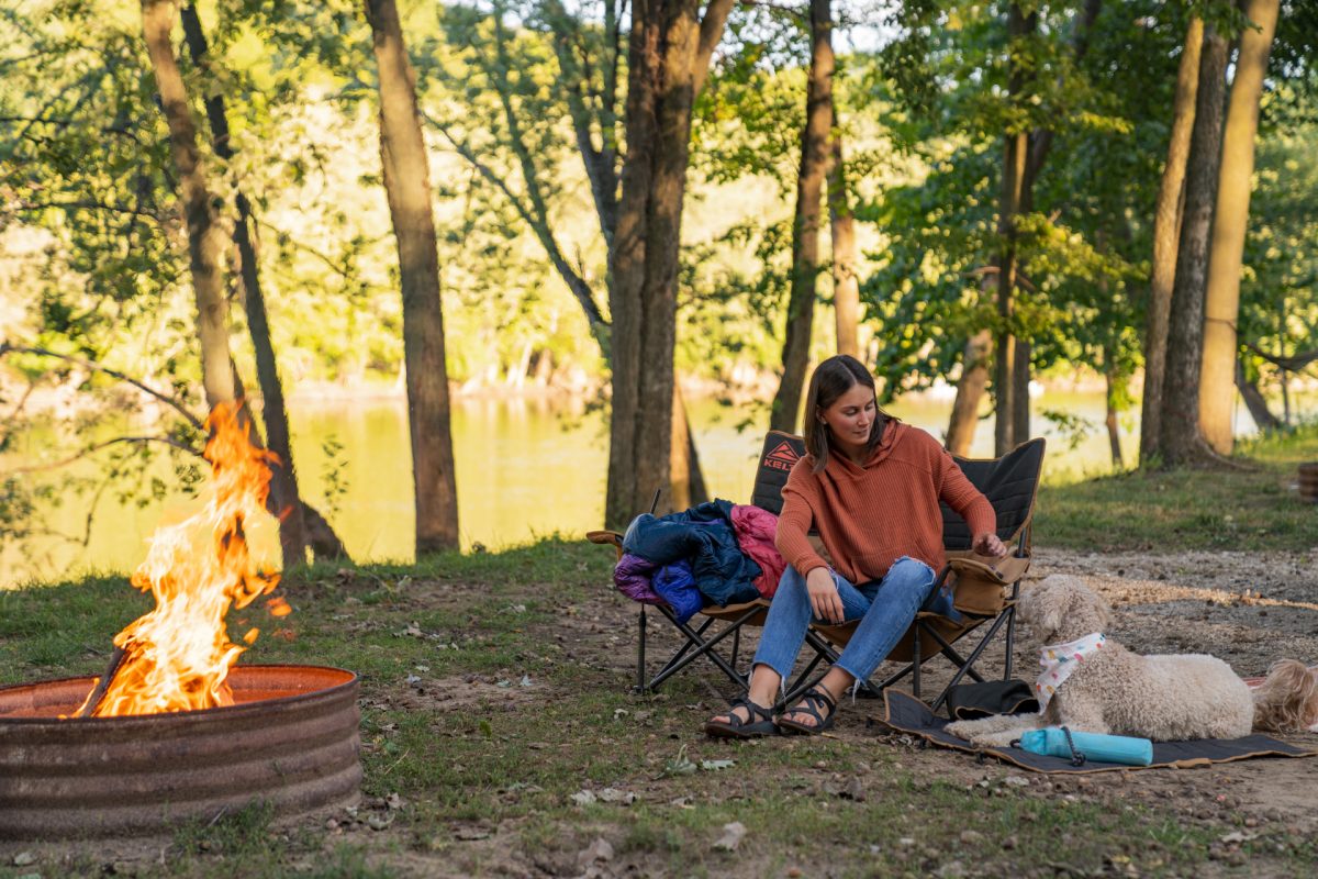 A woman with her dog while camping and sitting in front of a campfire.