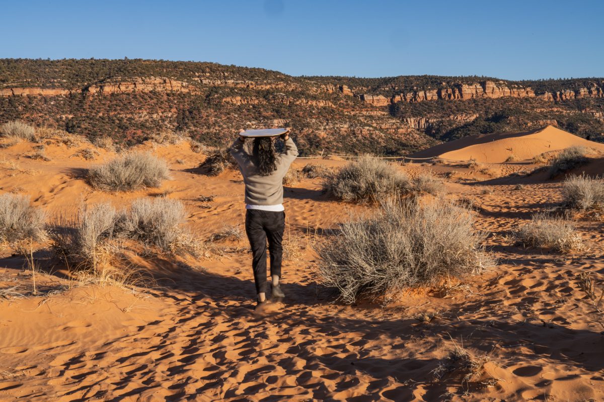 A person hikes out to the dunes at Coral Pink Sand Dunes State Park with a sand sled atop her head. Visiting Coral Pink Sand Dunes State Park is one of the best things to do in Kanab, Utah.