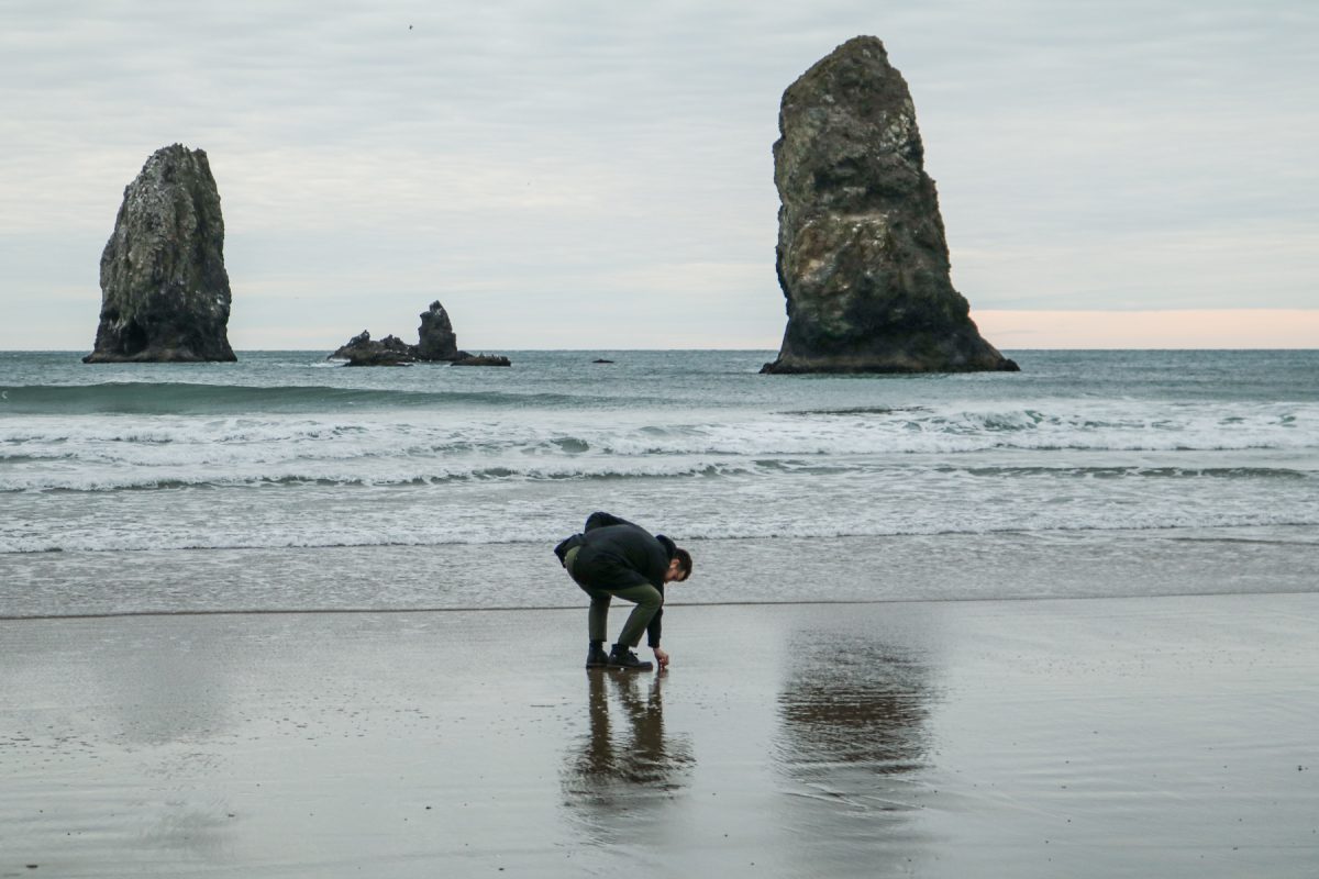 A man looking at rocks at Cannon Beach with the Needle sea stacks in the distance.