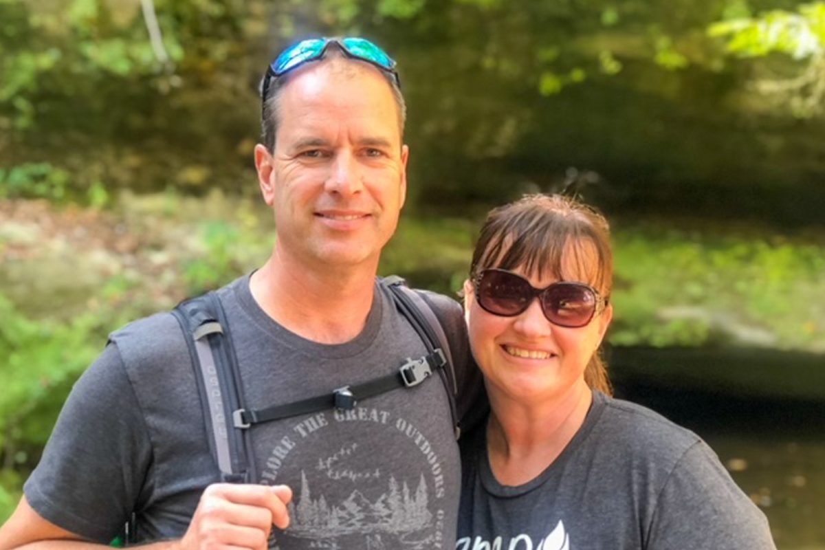 Two hikers smiling in front of a wooded area. 