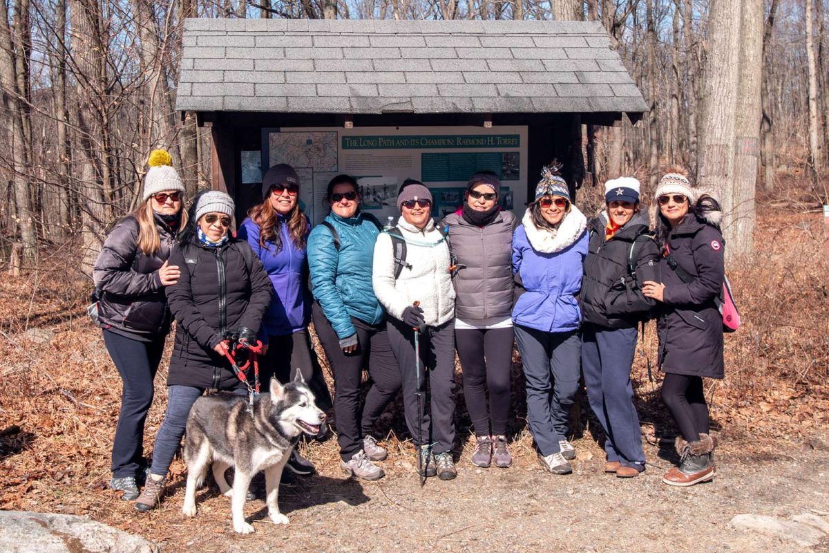 A group of hikers stands in front of a trail map sign dressed in winter gear. One hiker holds a leashed husky. 