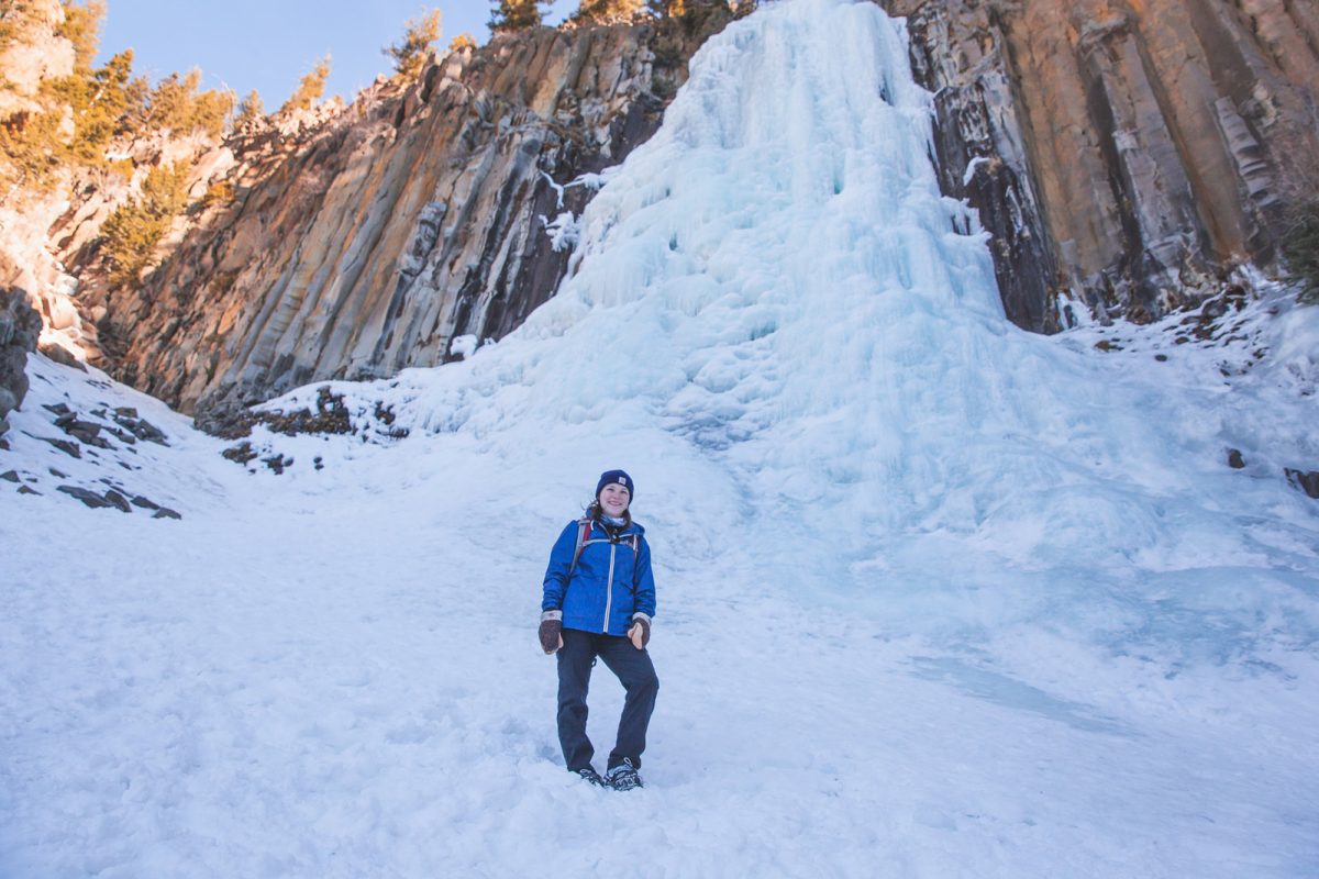 A person stands in front of a frozen waterfall dressed in winter hiking clothing and accessories. 