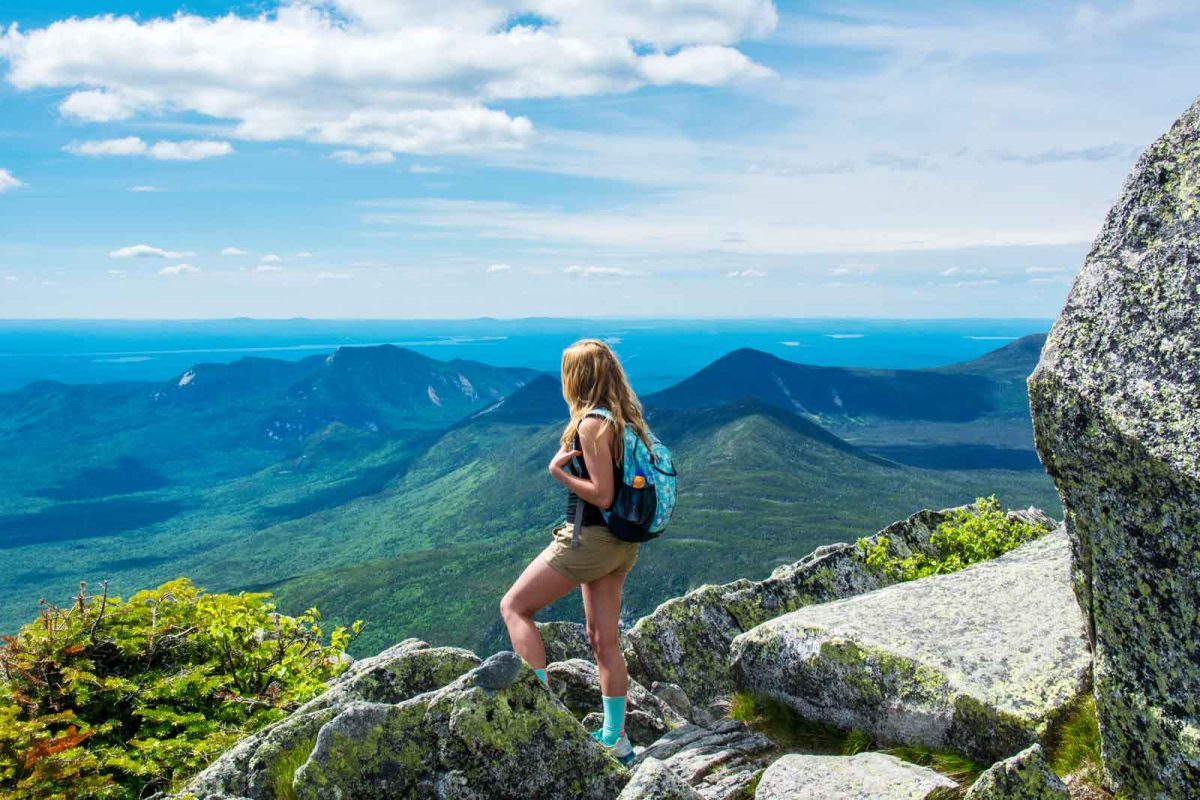 View from the top of Mt. Katahdin. A person dressed in a pack and shorts looks out over the view of green mountain tops in the distance. 