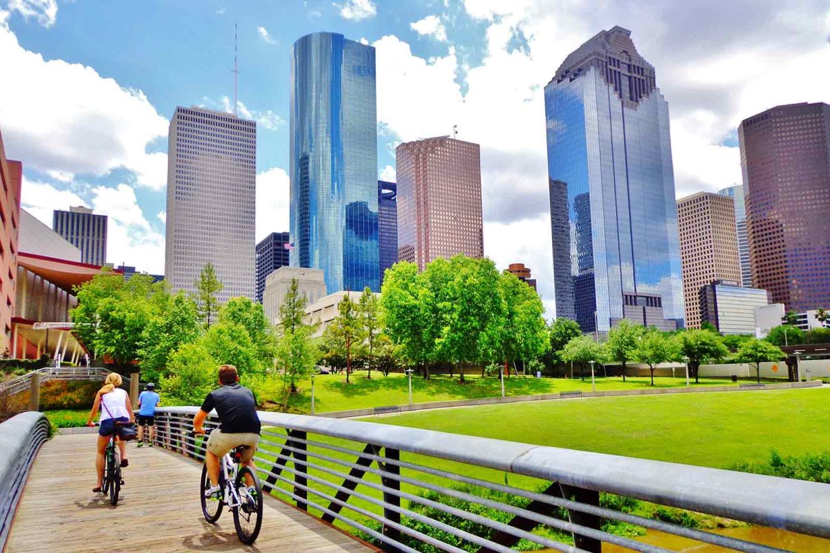 Cyclists cross a bridge in Buffalo Bayou with Houston skyscrapers visible in the distance.