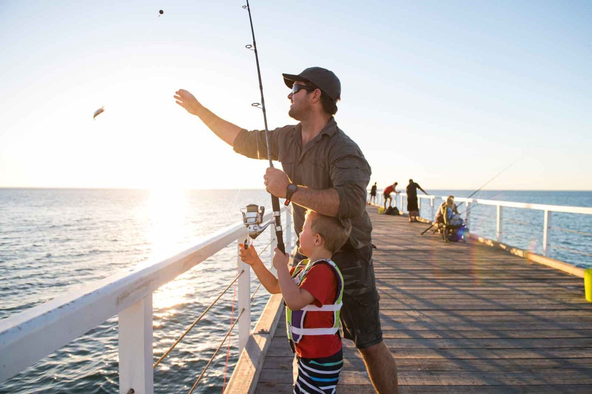 An adult fishes off of a pier with a young boy in a life vest. 