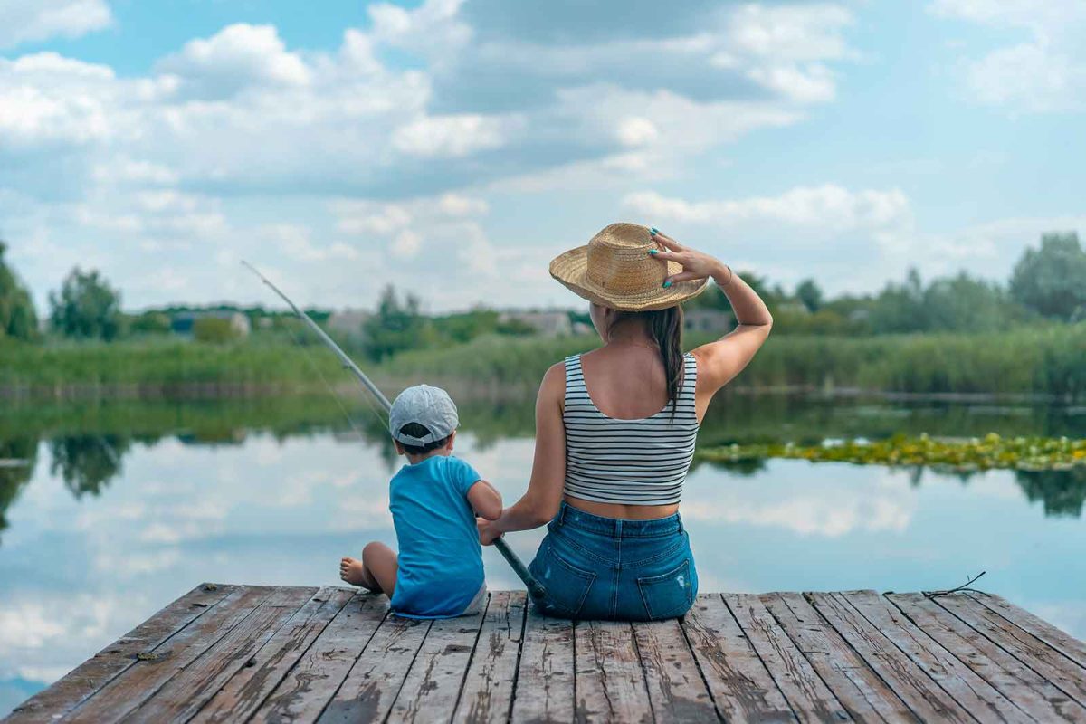 A woman in a hat sits on a dock and helps a young child hold a fishing pole. 