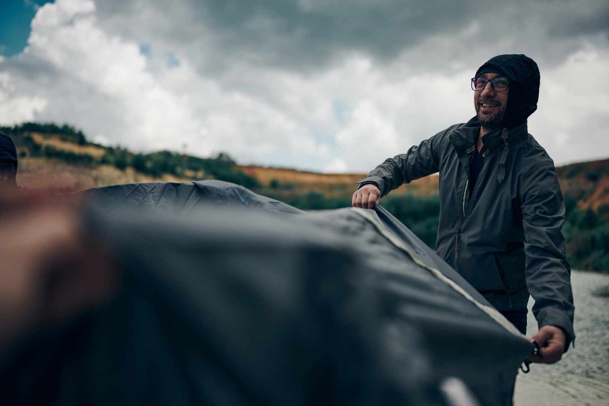 A man wearing a rain jacket helps to spread a tarp at a camping site in the rain. 