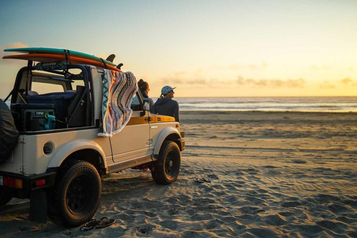 Two surfers lean on the front of a jeep parked on the sand, oceanside. 