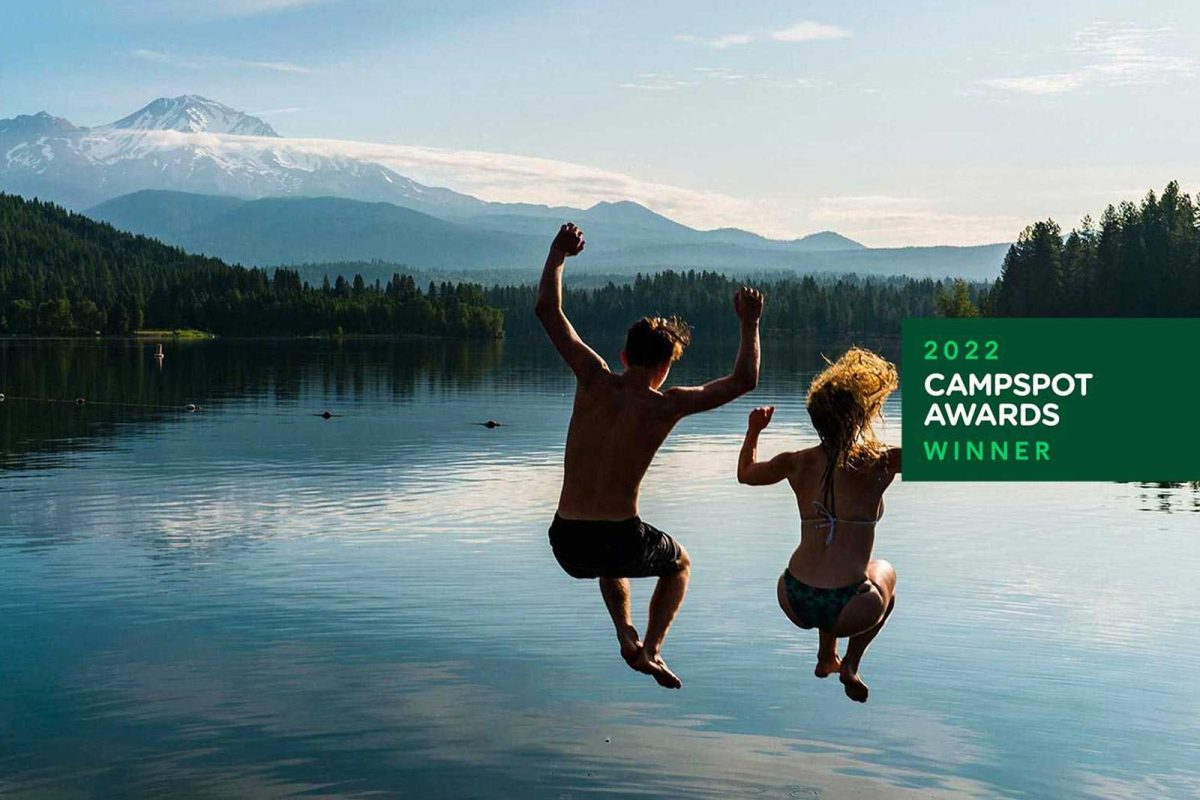 Two swimmers mid-air jumping into a lake with mountains and trees in the distance. 