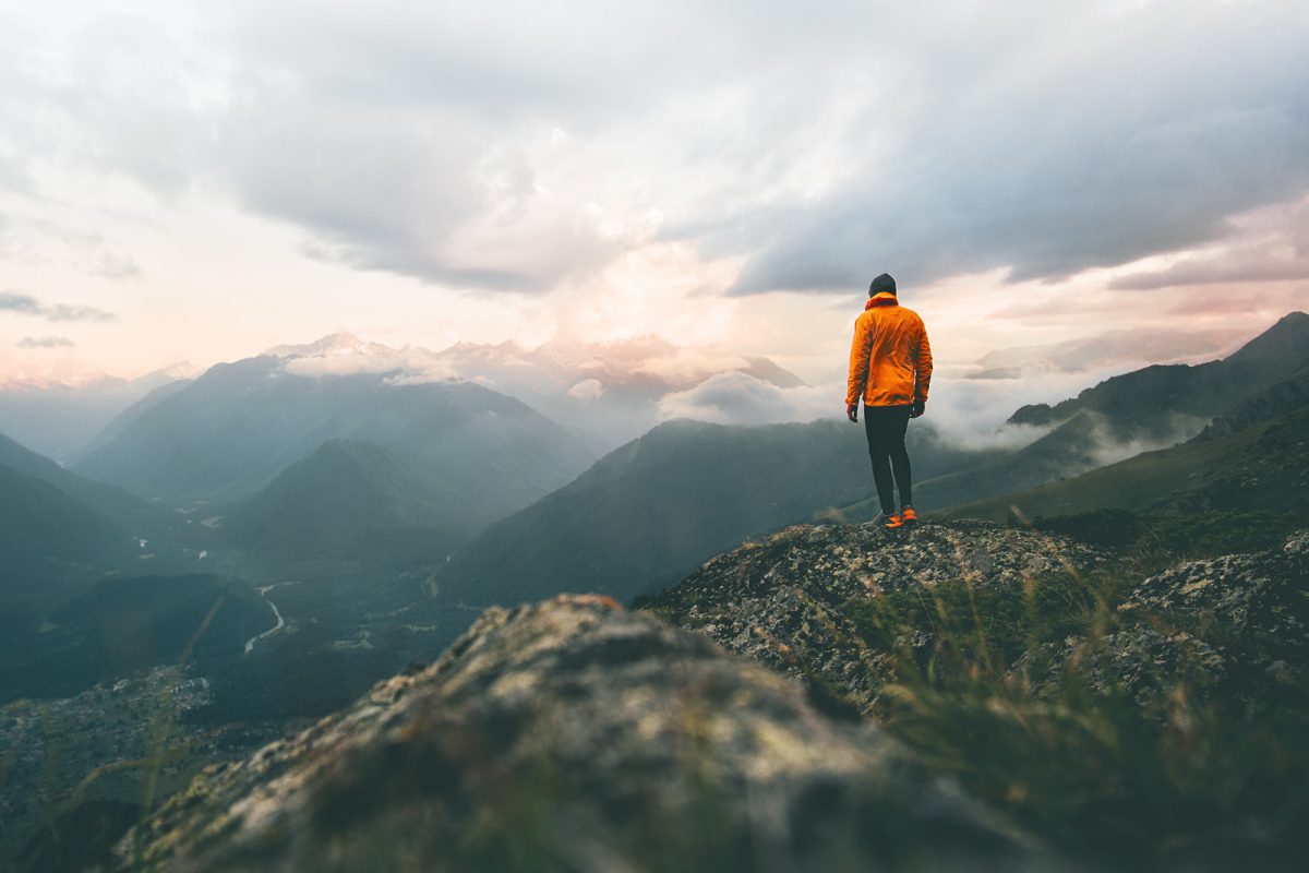 A trail runner in an orange jacket stares out at a mountain range from a vantage point on the trail. 