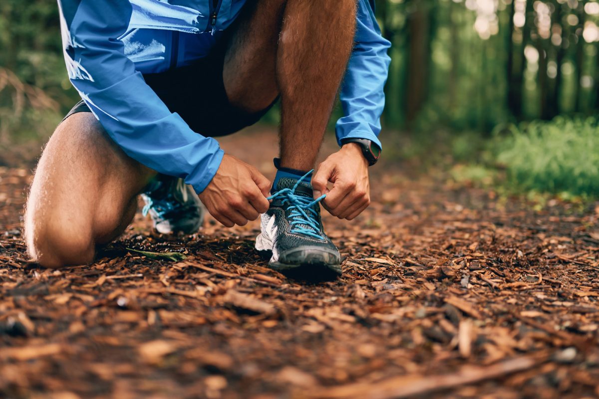 A runner stoops to tie their trail running shoes on a forest trail. 