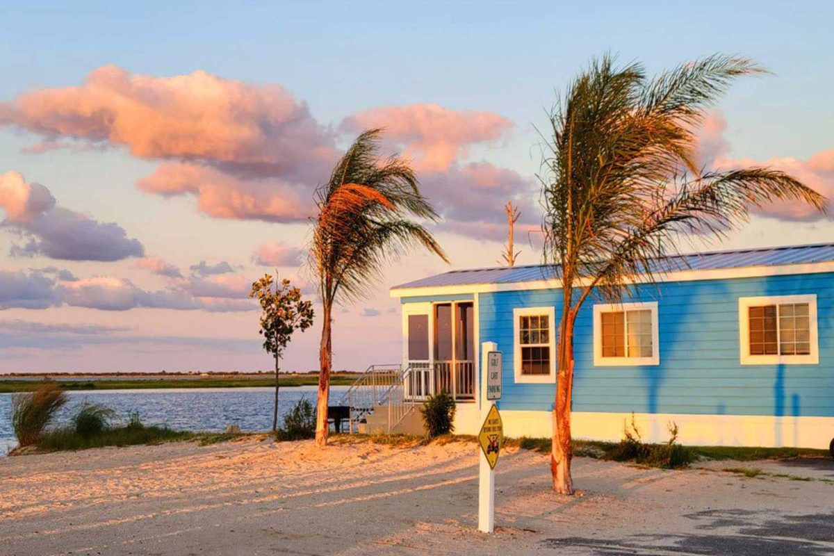 A blue camping cabin with palm trees at a campground on the water. 