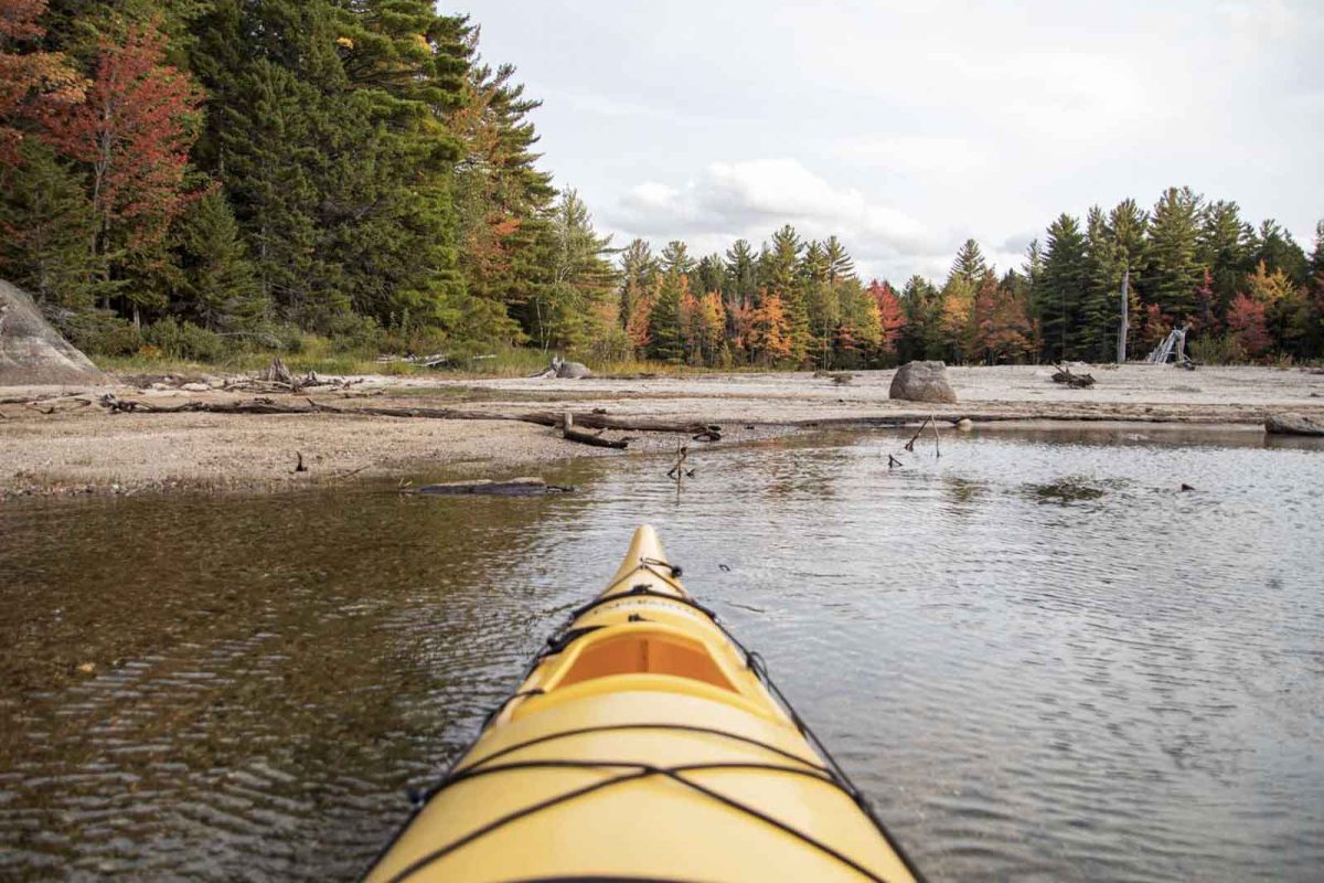 The front of a yellow kayak from the perspective of the kayaker on a body of water surrounded by trees in Maine. 