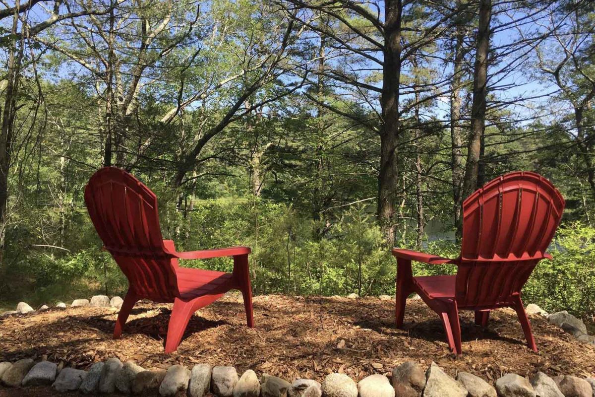 Two red Adirondack chairs overlook a body of water through the trees at a New England campground, offering the ideal spot for New England leaf peeping. 