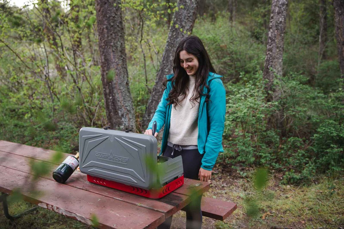 A woman cooking at a camp stove on a picnic table. 