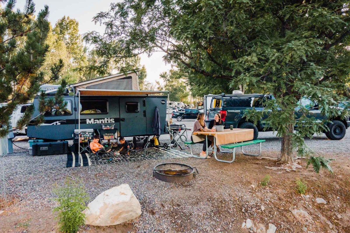 An image of an RV at a campground with a family spending time at a picnic table and under their awning. 