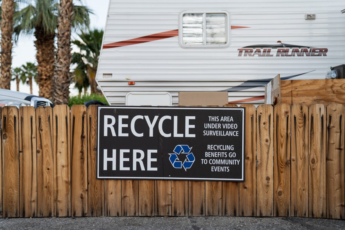 A sign on a fence reads, "recycle here" with an RV and palm trees in the background. 