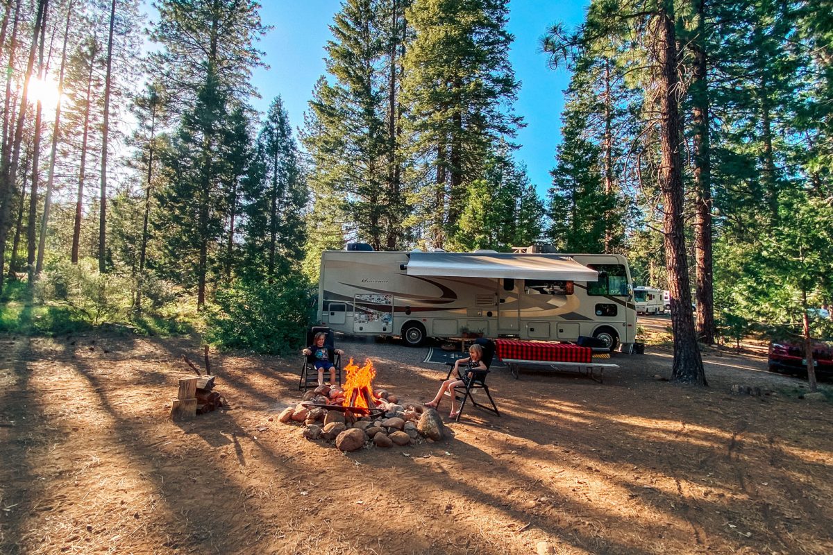 Two kids sit in lawn chairs beside a fire pit with an RV parked in the background. A picnic table is next to the RV and there are trees in the background. 