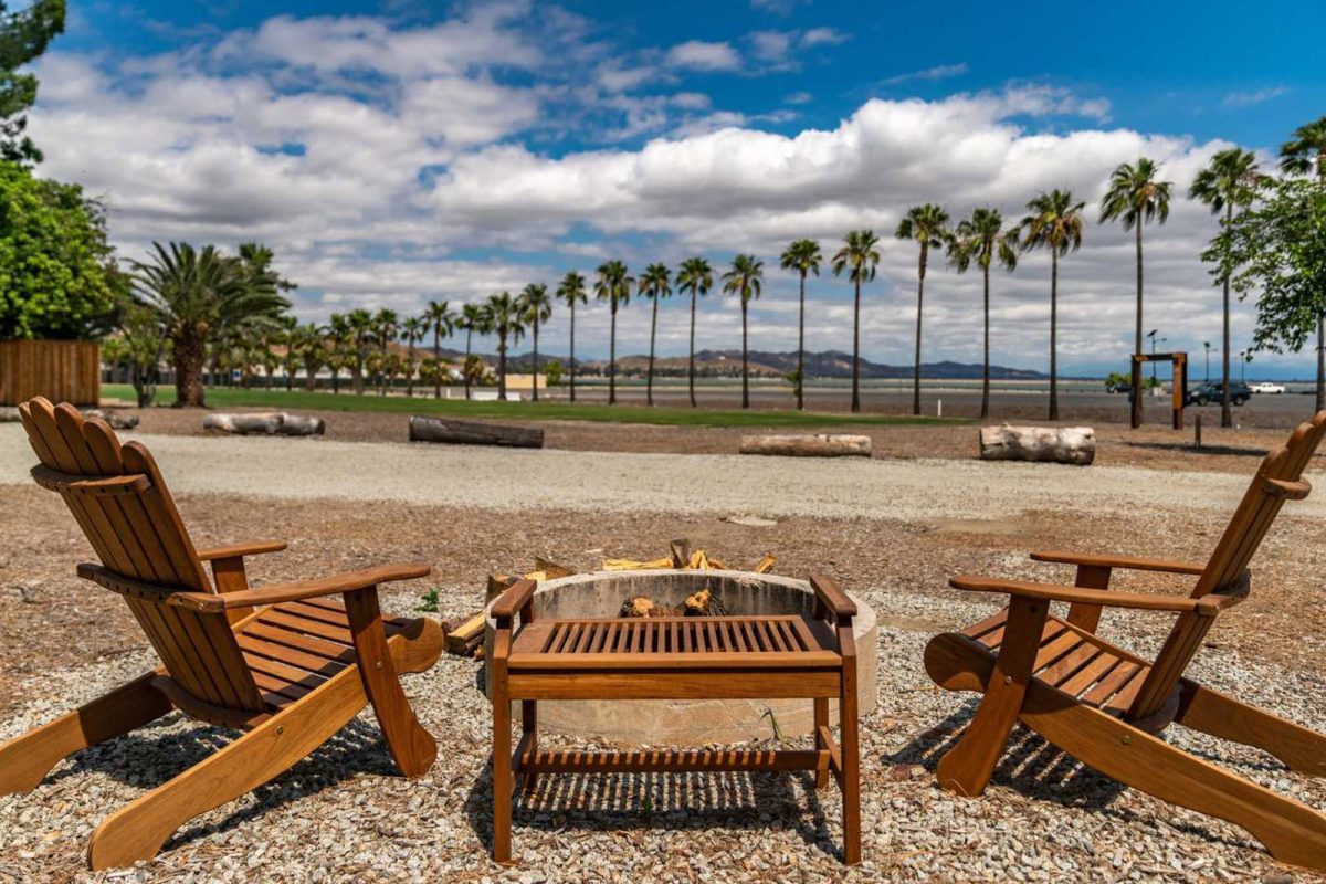 Two Adirondack chairs on either side of a fire pit with palm trees in the distance. 