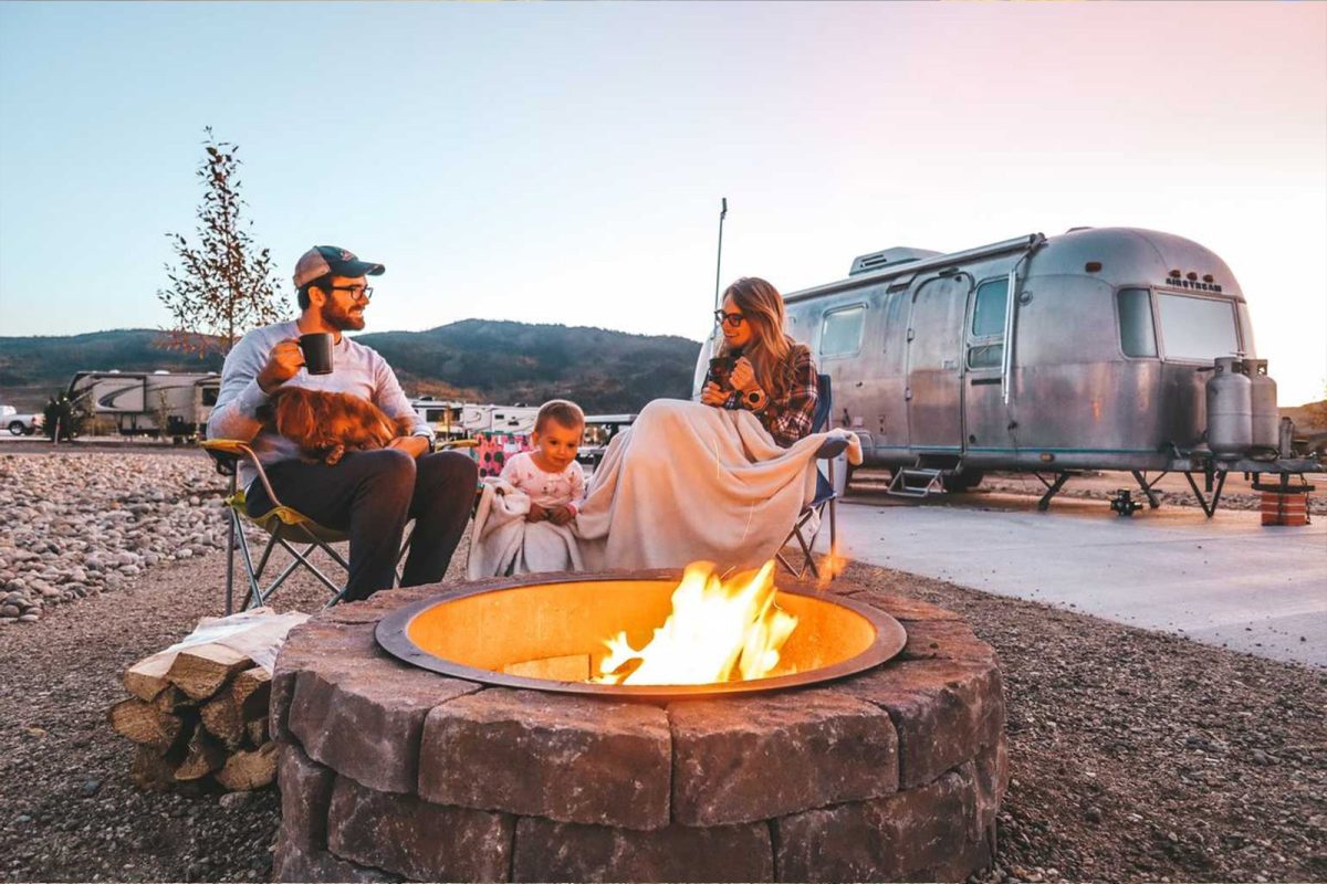 A family sits around a campfire with an Airstream in the background at Sun Outdoors Rocky Mountains, one of the most popular campgrounds for stargazing. 