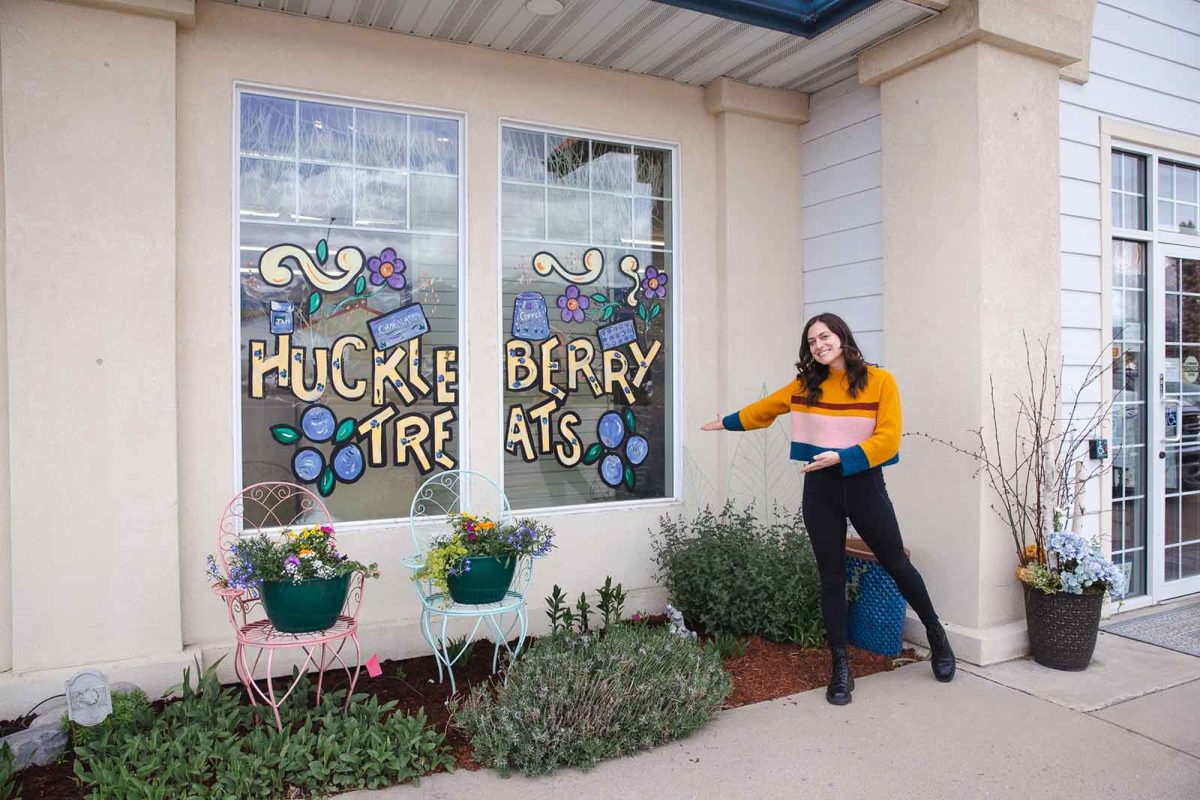 A woman in a yellow sweater stands in front of store window decorated with the words "Huckleberry Treats." 