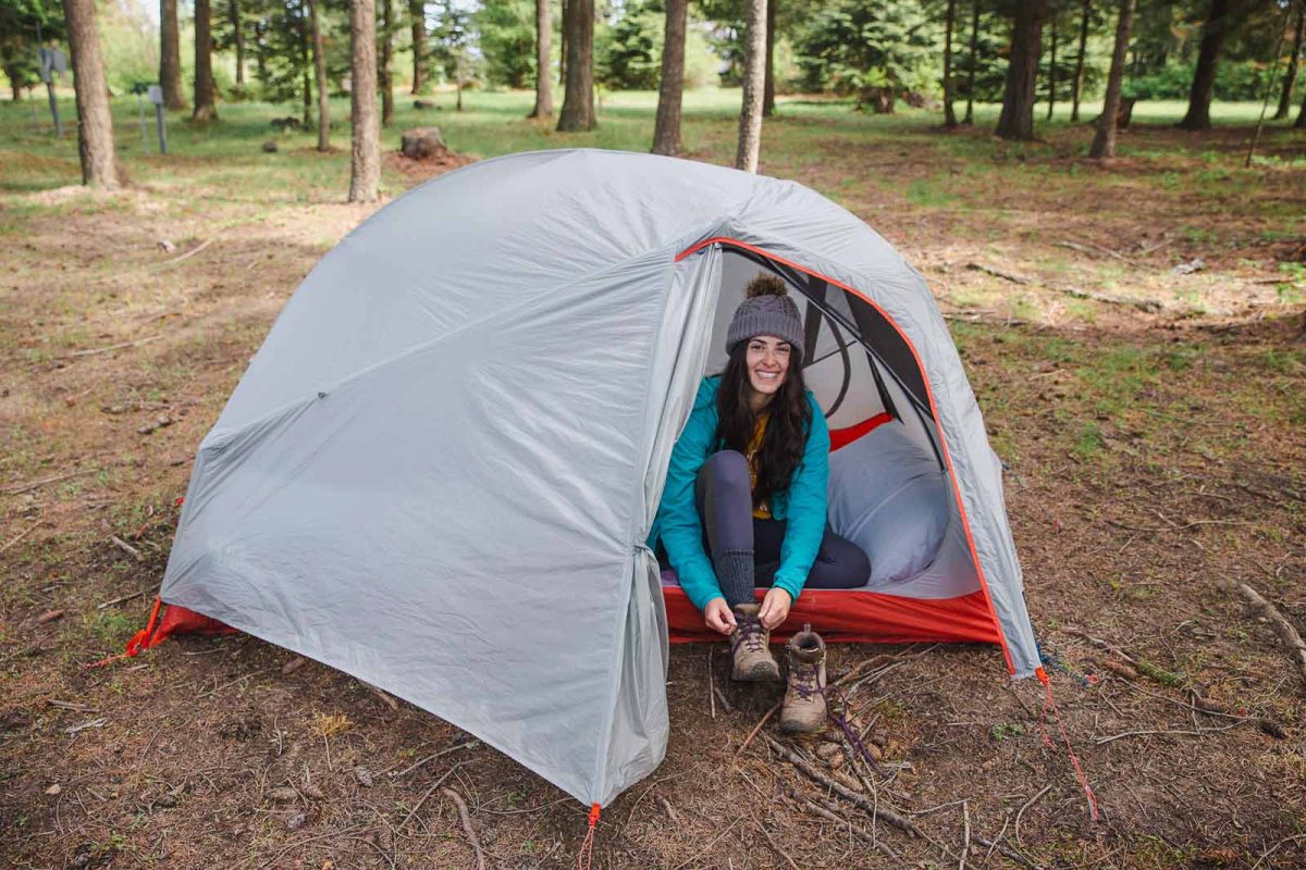 A woman laces her hiking books from inside her tent with trees in the background. 