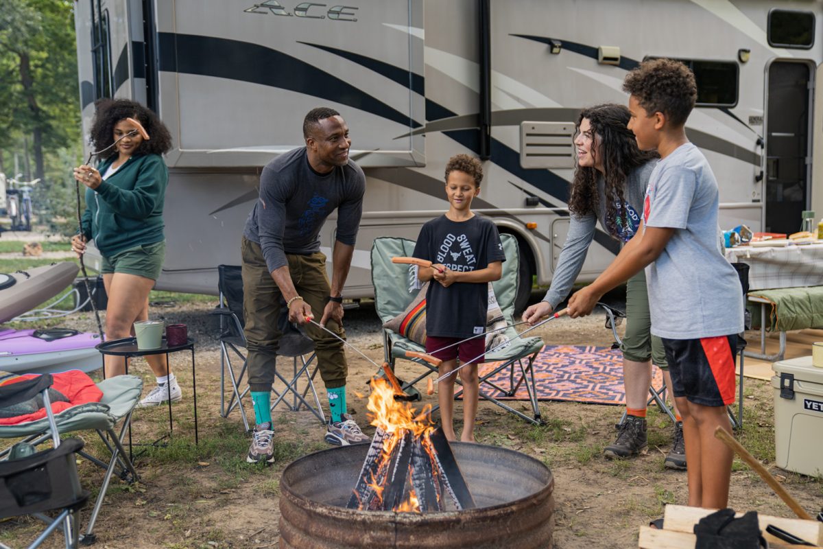 A group of campers roast hot dogs over a fire in front of an RV. 