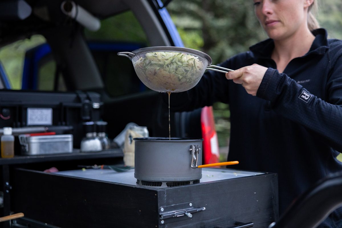 A woman strains grated zucchini and potato through a mesh strainer over a pot on a camp stove. 