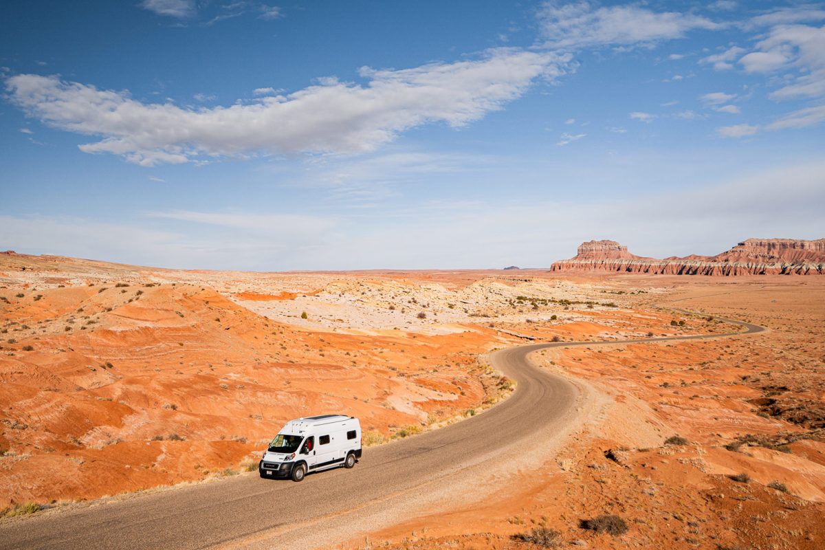 An RV on a desert road with orange sand and rock formations in the background. 