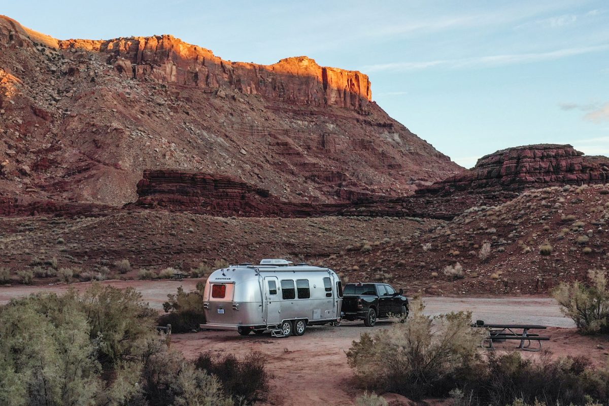 A black truck tows an airstream trailer in the desert with rock formations in the background. 