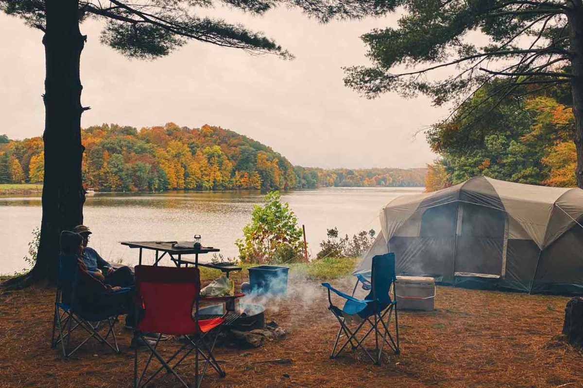 A tent and fire pit and camping chairs next to a body of water with fall foliage in the background 