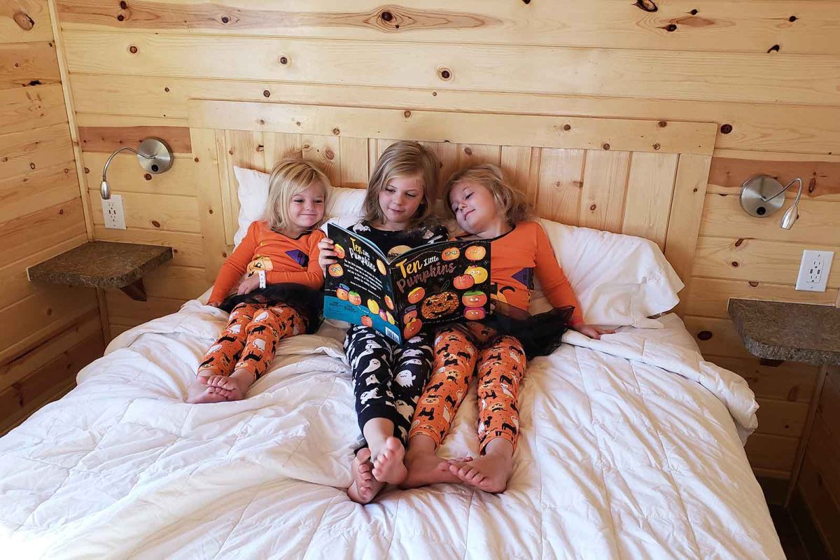 three children in Halloween costumes reading a Halloween-themed book on bed