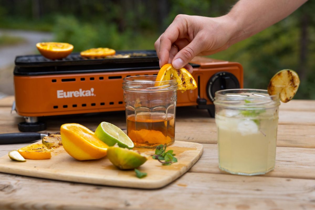 Hand places a grilled orange slice on a camping cocktail glass. 