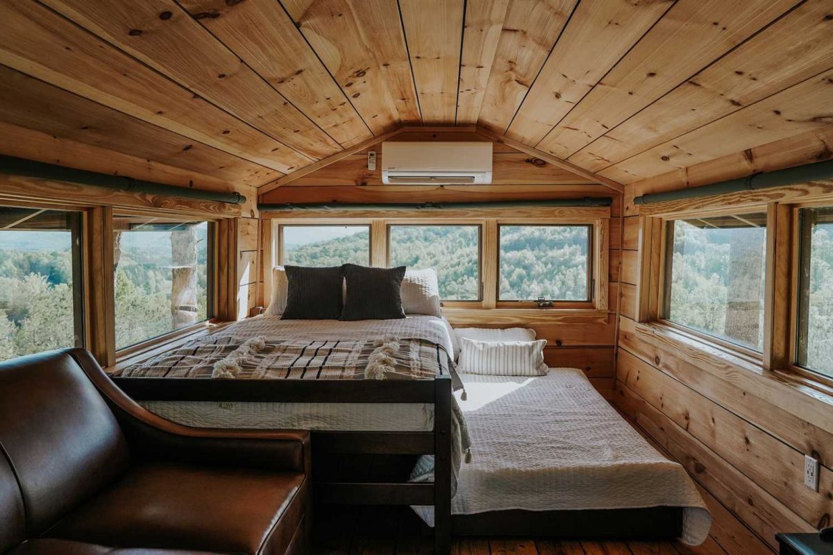 A bed in a lofted cabin with windows overlooking the wooded hillside below 