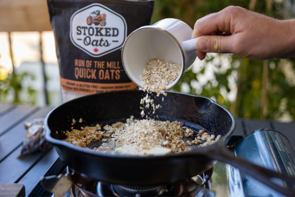 A hand pours oats from a coffee mug into a skillet on a camp stove to make maple apple crisp 