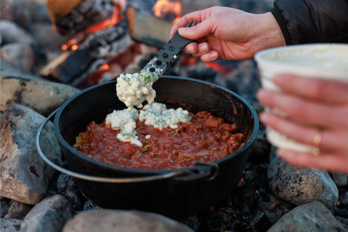 person layering ricotta cheese onto dutch oven lasagna camping recipe