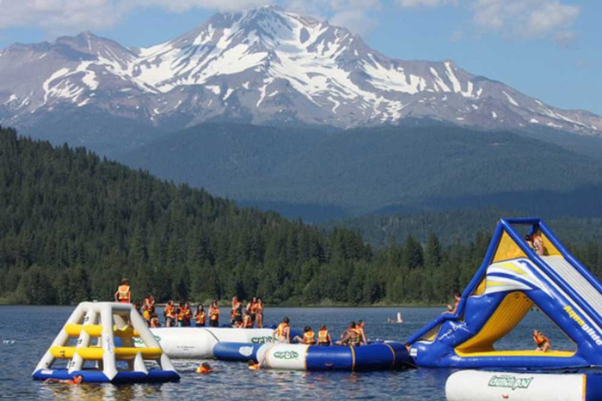 A large snow-capped mountain looms over hills studded with evergreens with campers enjoying an inflatable waterpark in a lake in the foreground. 