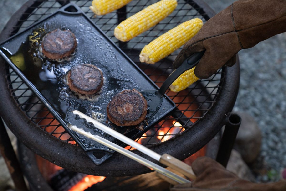 camper cooking meat patties and grilling corn over campfire