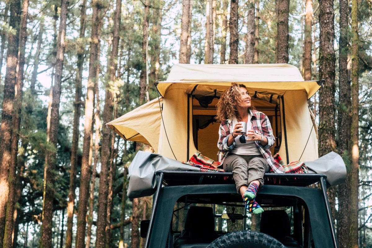 woman relaxes while camping and drinking coffee on top of her car